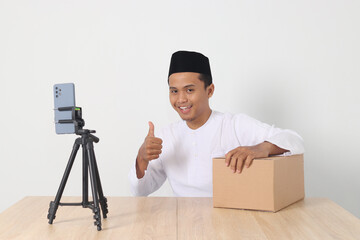 Portrait of excited Asian muslim man in koko shirt with skullcap promoting his product on live streaming session. Online shopping with smartphone concept. Isolated image on white background