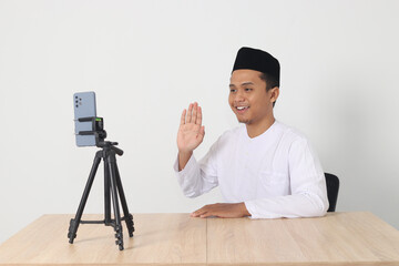 Portrait of excited Asian muslim influencer in koko shirt with skullcap saying hi, waving hand to...