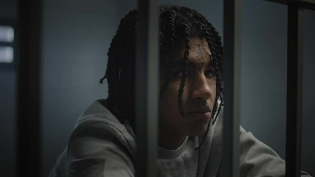 Angry African American teenage prisoner with face tattoos stands in prison cell in jail and looks at camera. Young criminal serves imprisonment term for crime. Juvenile detention center. Portrait.