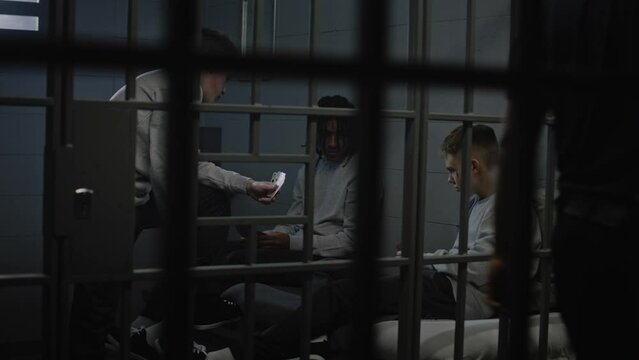 Three multiethnic teenage prisoners play cards in prison cell. Young criminals serve imprisonment term for crimes in jail. Juvenile detention center or correctional facility. View through metal bars.