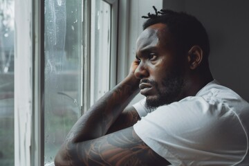 depressed african american man in white t-shirt with tattoo on arms looking through window