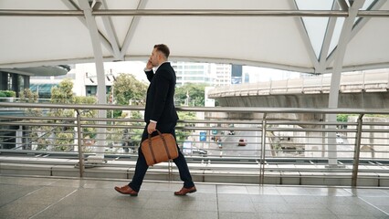 Smart caucasian businessman calling his colleague to plan financial strategy while walking to workplace. Side view of manager using mobile phone to communicate with marketing team. Lifestyle. Urbane.