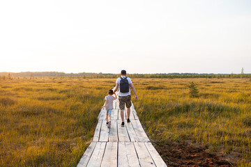 Man with child  walking in wildlife national park at sunset. Wooden boardwalk through swamp in late summer. Concept of ecotourism, hiking, vacation, travel with children. Selective focus.