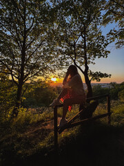 City overlook: woman sits on fence, captivated by sunset.