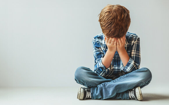 A little child or boy sits with his head bowed and crying, his hands covering his face. Sitting sad and disappointed on the floor and next to the wall. Copy space.