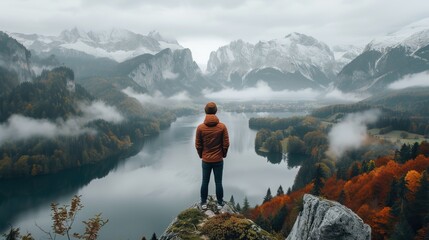 Person in orange jacket standing on cliff overlooking misty mountain lake in autumn. - obrazy, fototapety, plakaty
