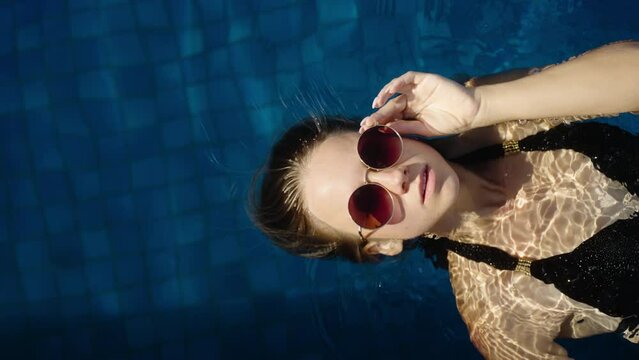 Portrait of woman in sunglasses floating and relaxing in a pool. Total relax and chilling in the water, vertical video. Summer vacation tour in pool private villa or hotel