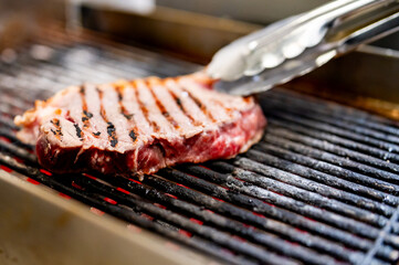 juicy steak grilling with perfect sear marks, held by tongs over a hot grill. The steak’s distinct grill marks suggest it’s being cooked on a grill with parallel bars.