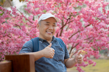 Asian middle-aged man in white cap and has backpack standing and thumping up in front of sakura flower tree, soft focus, happiness of middle-aged people and a single man around the world concept. © Sophon_Nawit