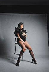 Fototapeta na wymiar Attractive pregnant woman in black bodysuit, nylon stockings and high heels posing in studio. The concept of stylish motherhood.