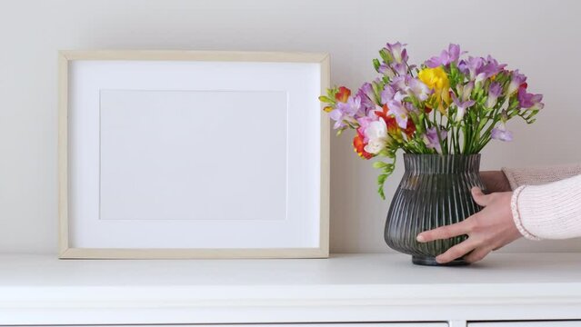Female hands puts flowers in a vase. Wooden frame mockup in white room interior, empty mock-up with copy space. Light frame with a vase in the interior for design presentation.