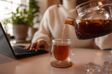 The girl works at the computer and drinks tea in cafe. Close-up of female hands pouring tea from the teapot