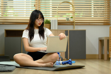 Young woman sitting on on exercise mat and using laptop computer for watching online tutorial