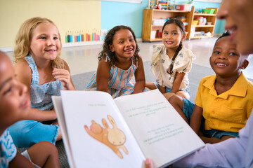Female Primary Or Elementary School Teacher Reads Story To Multi-Cultural Class Seated In Classroom