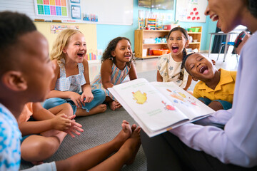 Female Primary Or Elementary School Teacher Reads Story To Multi-Cultural Class Seated In Classroom