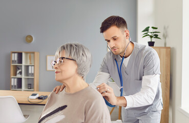 Obraz premium Young male nurse attending to senior woman patient. Man doctor in scrubs uses stethoscope to examine heart and lungs of old lady who came to clinic for routine medical checkup