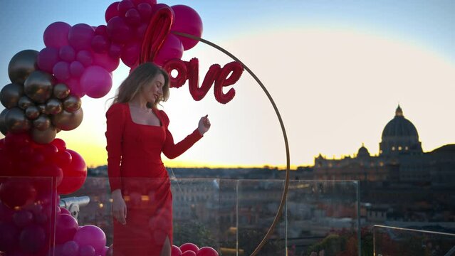 Beautiful young woman in red dress dancing at sunset near the red and pink balloon arch spelling out the word "love". Vatican city in background. Rome, Italy