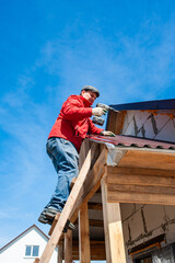 Fototapeta na wymiar A worker builds a roof in a house while standing on a wooden ladder. Blue sky