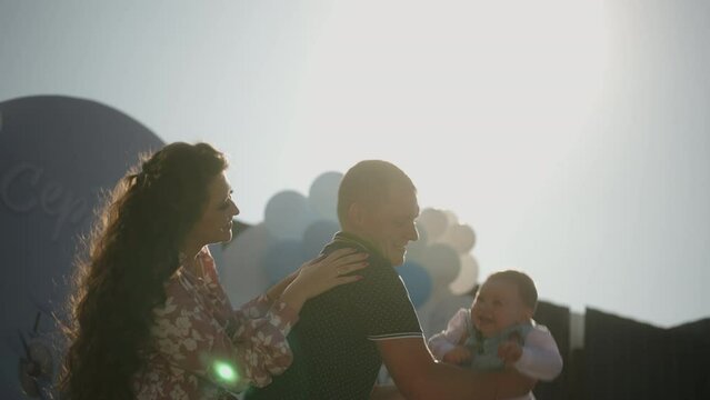 Family celebrate birthday party of son, father dad tosses up baby in rays sunset, smile laugh kiss. Concept of happy memories of childhood, party decoration on background 