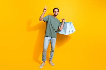 Full length photo of friendly funny guy wear khaki t-shirt holding shoppers showing v-sign isolated yellow color background