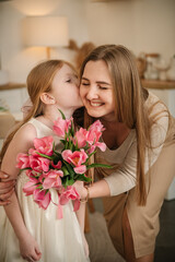 Daughter kisses and hugs mom. Happy family holiday. Cute little child girl greets smiling mother in kitchen. Beautiful kid gives mother big bouquet of tulip flowers and makes surprise for Mother's Day