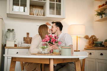 Romantic female kisses male, spending time together. Man congratulate woman and give bouquet of flowers on Women's day. Loving couple having conversation and drinking coffee in morning in kitchen.