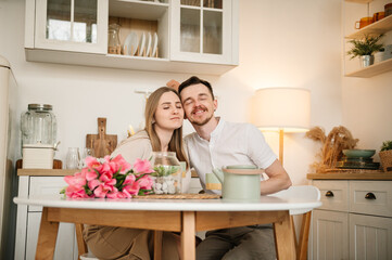 Fototapeta na wymiar Loving couple having conversation and drinking coffee in morning in kitchen. Romantic male hug female, spending time together. Man congratulate woman and give bouquet of flowers on Women's day.