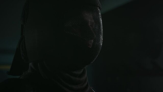 Side closeup of Caucasian male foil fencer putting on black metal mesh face mask before fight in dark hall