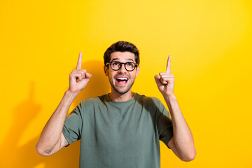 Photo of cheerful excited guy wear khaki t-shirt spectacles showing two fingers looking up empty space isolated yellow color background - obrazy, fototapety, plakaty