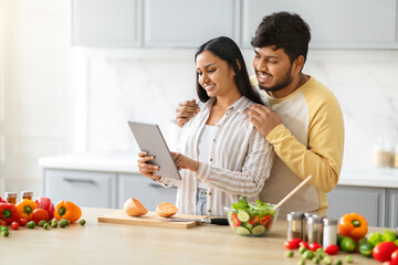 Happy loving young indian spouses using digital tablet while cooking