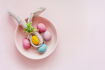 Ein Osterei mit einer Stoffserviette als Hasenohren und bunte Eier in einer Schüssel. Draufsicht,...