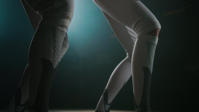 Ground level tilt footage of anonymous fencing instructor teaching starting pose to fencer during first workout in dingy hall