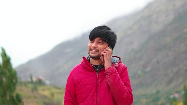 Portrait of a happy young Indian man wearing jacket talking on the mobile phone in front of the mountains at Manal in Himachal Pradesh, India. Happy man talks on the phone. Happiness due to call.
