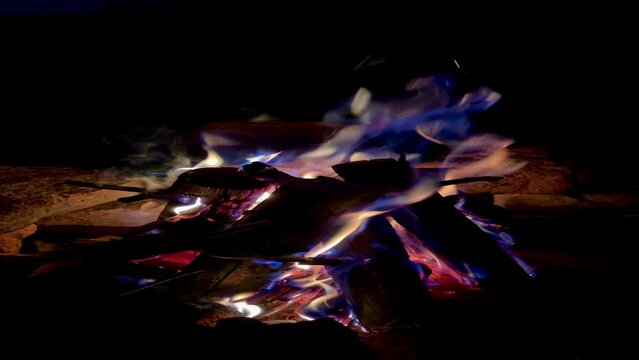 Close-up slow motion of a burning campfire at night, an open-air fire pit, firewood in flames, sparks and smoke, bonfire, outdoor camping, relaxation, warmth, yellow-blue flame