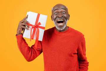 Ecstatic senior black man in red sweater holding a gift with a red ribbon to his ear