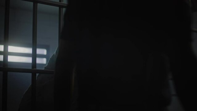Upset African American teenager with face tattoos stands in prison cell in jail or youth detention center leaning on metal bars. Prison officer passes by young criminal or prisoner in the foreground.