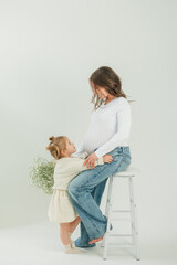 Beautiful young pregnant woman with bouquet of spring flowers with her little daughter 3 years old on white background. Stylish model with belly in white bodysuit and jeans. Mother's Day.