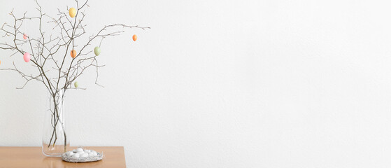 Vase with tree branches and Easter eggs in nest on table near white wall - obrazy, fototapety, plakaty
