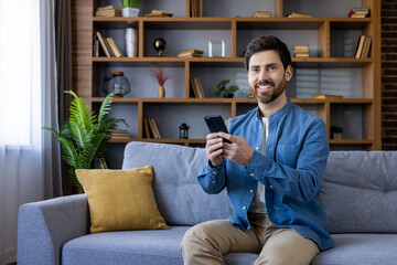 Friendly man sitting on couch with smartphone at home