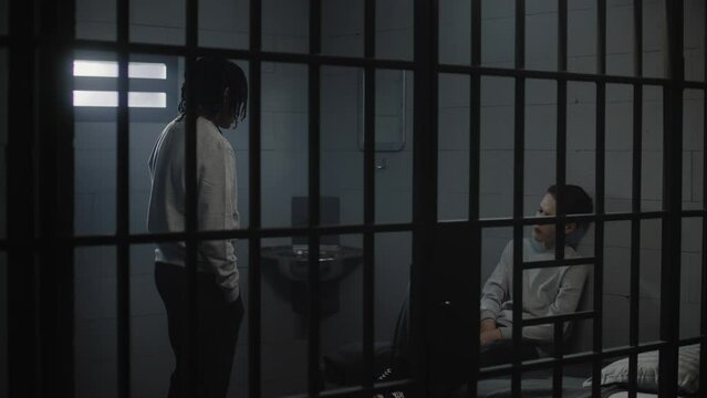 Multiethnic teenage prisoners talk in prison cell, serve imprisonment term for crime in jail or youth detention center. African American jailer passes by young criminals or inmates in the foreground.
