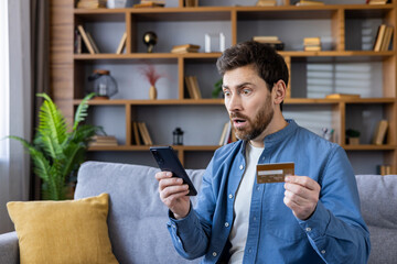 Surprised man holding smartphone and credit card at home