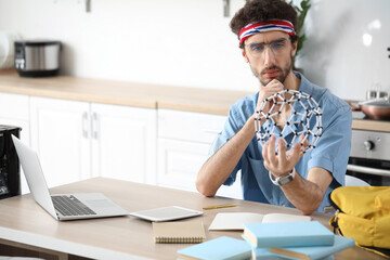 Thoughtful male student with molecular model studying at table in kitchen