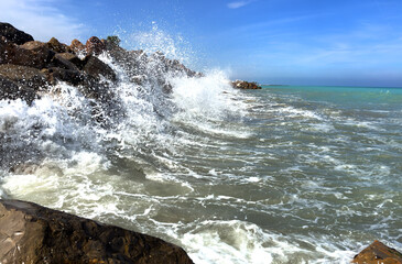 Waves splash on stones in sea. Breakwater with sea waves. Wave at stone pier. Wave in a storm breaks on stones at pier. Waves splash on stones at coast beach. Sea waves breaking on rocks in ocean.