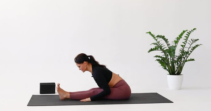 Athletic woman performs stretching exercise for back and leg muscles, working out in black sportswear while sitting on a mat in a light room