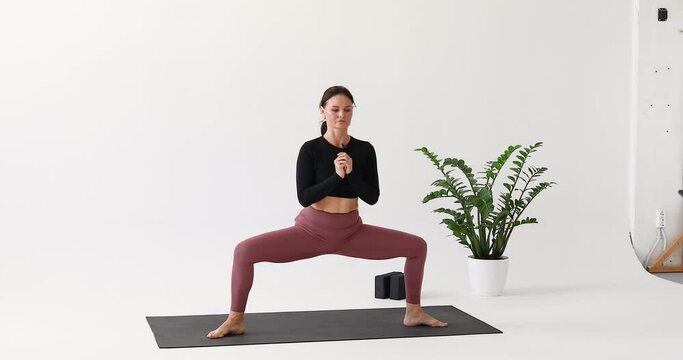 A woman moves from Rudrasana to Virabhadrasana 1, warrior pose with knee touching the floor, exercising in sportswear while standing on a mat in a bright room