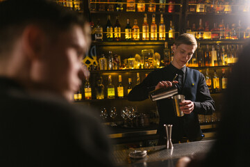 Friends sit at bar counter waiting for drinks from bartender. Male specialist in work uniform makes...