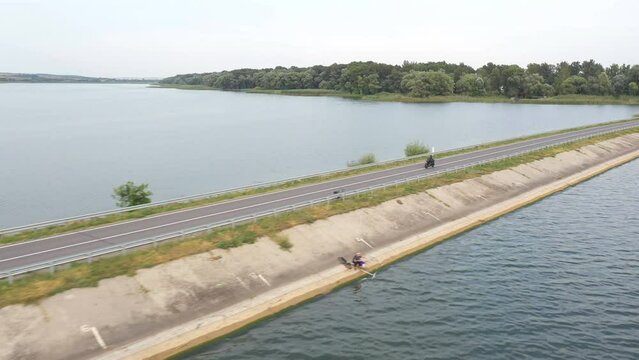 Aerial shot of motorcyclist racing his motorcycle through road near lake. Man drive bike at dam route on summer day. Flying over biker ride on modern sport motorbike through bridge of river. Top view