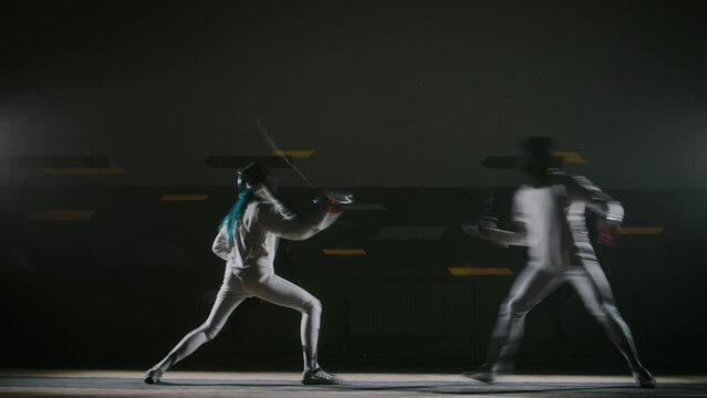 Side full timelepse of two professional fencers fighting on rapiers during duel in dark hall
