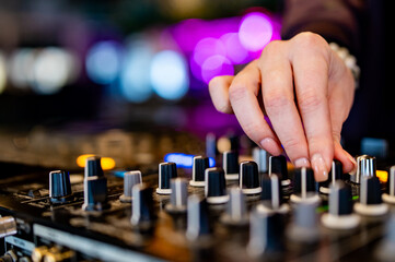 close-up view of a DJ’s hand adjusting knobs on a sound mixer. Various knobs and sliders are visible, indicating different channels and controls for audio mixing. The DJ’s hand is in focus