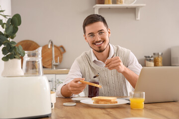 Fototapeta na wymiar Young man with laptop having tasty jam toasts for breakfast in kitchen